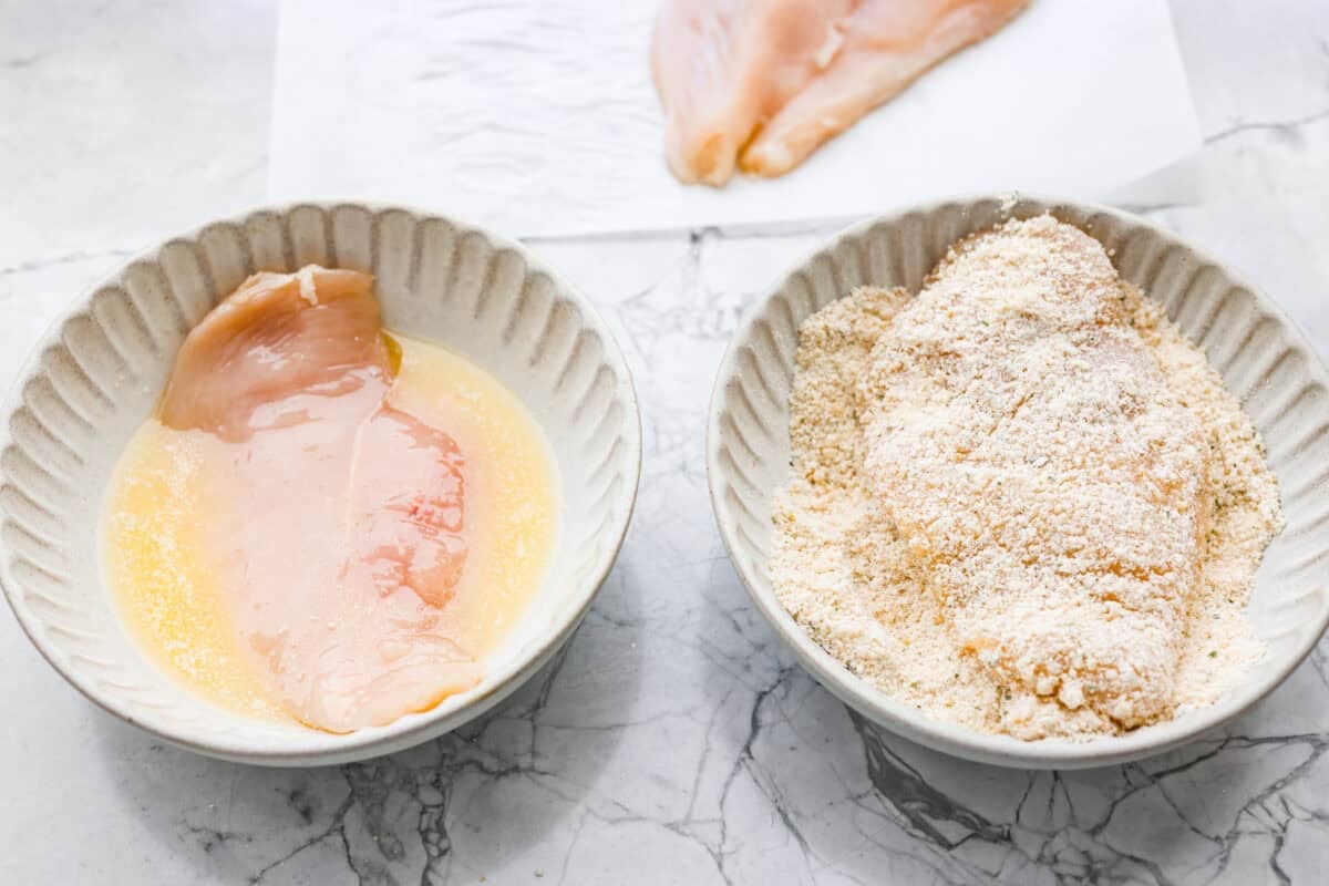 Chicken being dredged in egg wash and bread crumbs in separate bowls. 