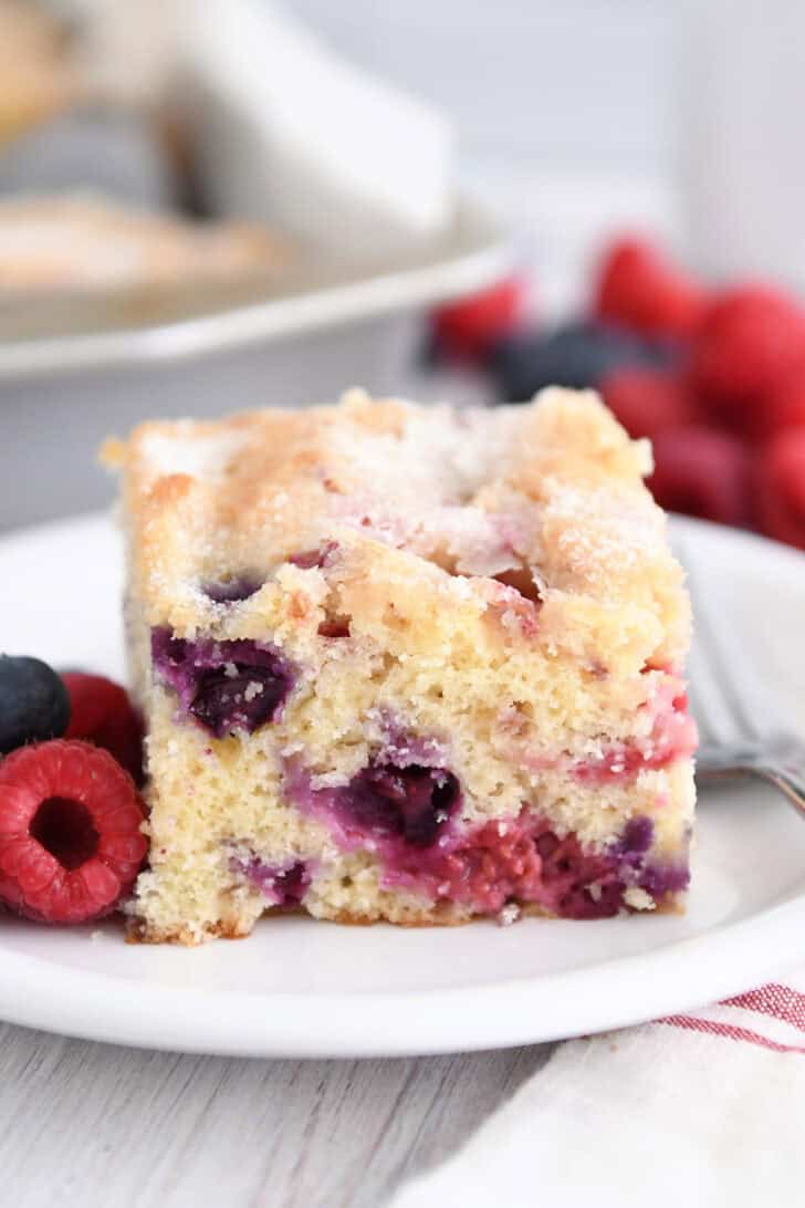 Square of cake with blueberries and raspberries on white plate.