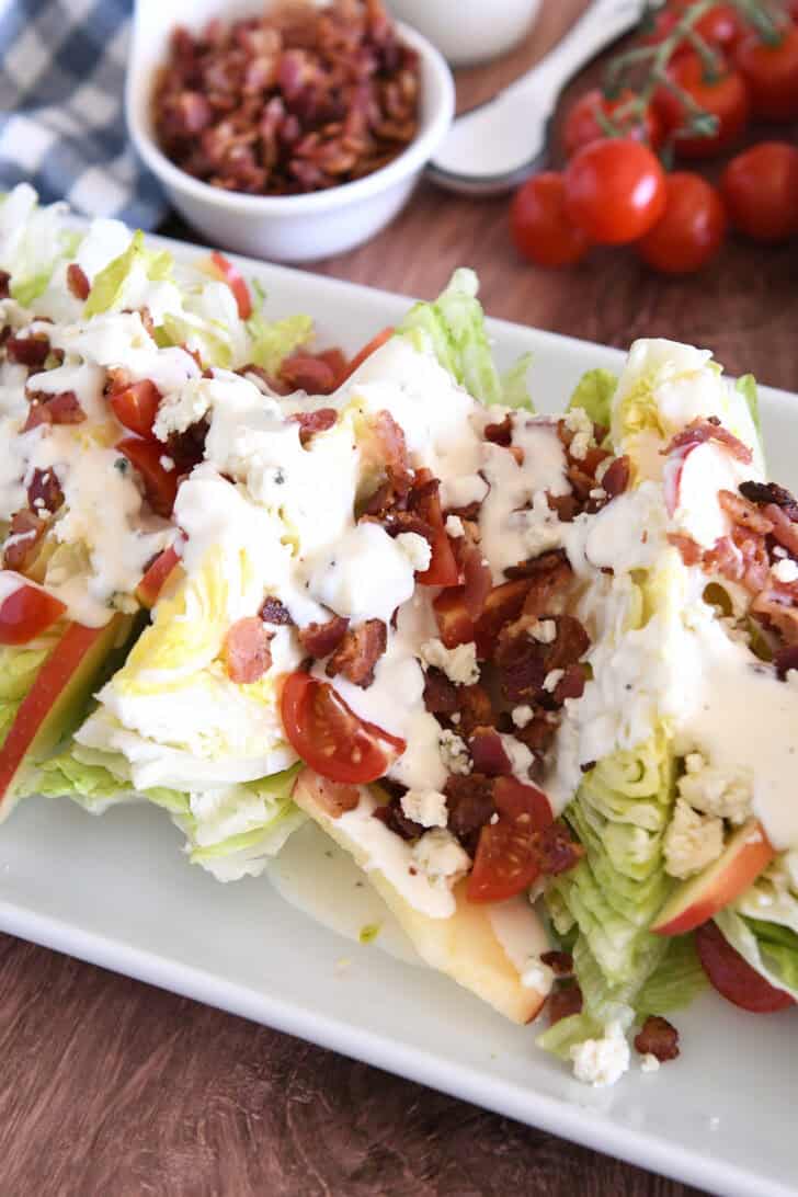 White platter with iceberg wedges, blue cheese crumbles, blue cheese dressing, tomatoes, bacon, and sliced apples.