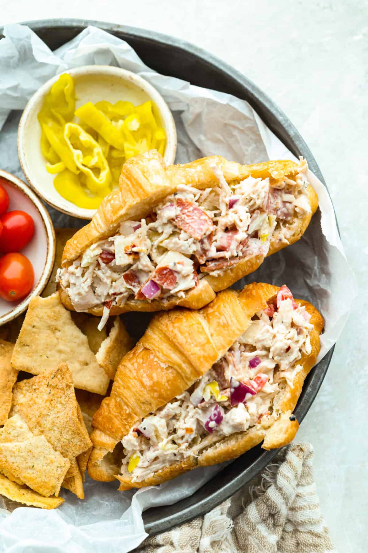 Platter of grinder chicken salad sandwiches next to pita chips and banana peppers. 