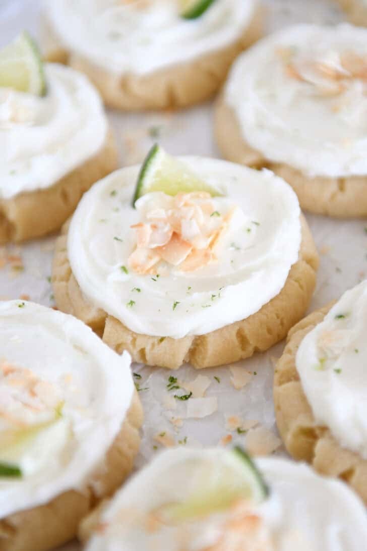 Lots of frosted coconut lime sugar cookies topped with toasted coconut and a fresh lime wedge on white parchment paper.