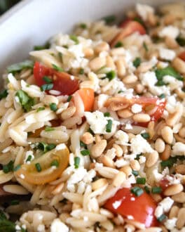 A wooden spoon taking a scoop of cooked orzo salad with tomatoes, pine nuts, and basil out of a white casserole dish.