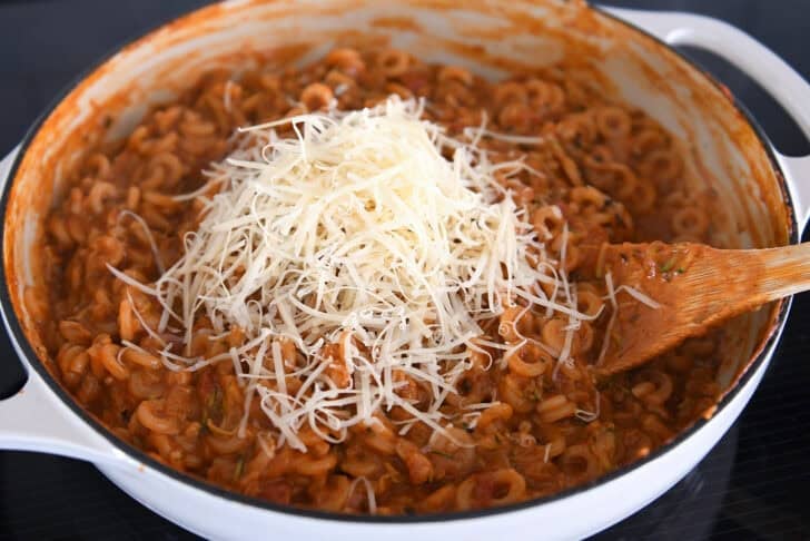 Adding freshly grated Parmesan to cooked pasta in red sauce with shredded zucchini in deep skillet.