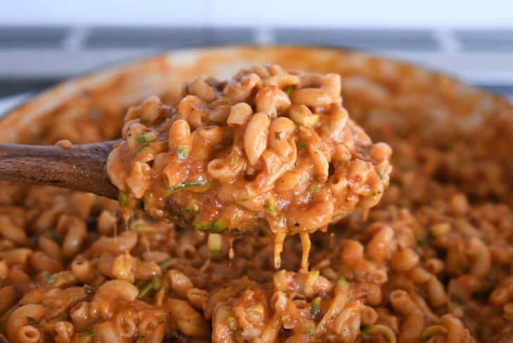Spoon scooping up serving of saucy pasta with shredded zucchini in skillet.