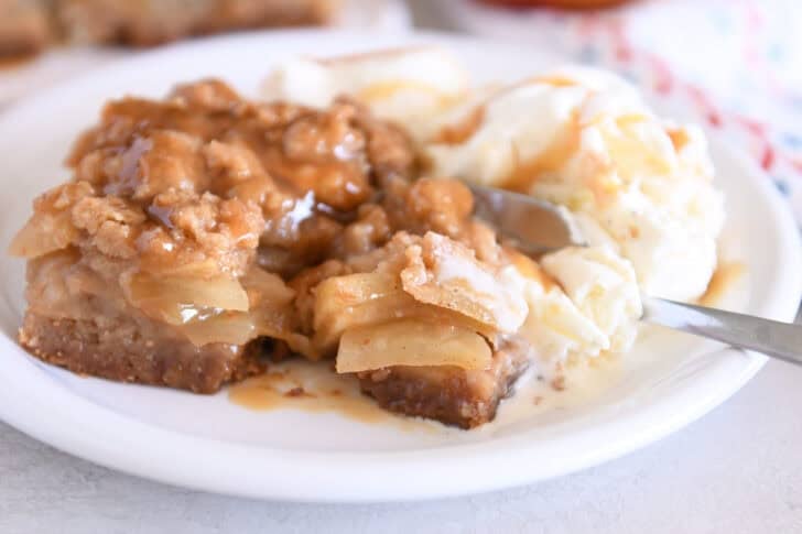 Fork scooping bite of caramel apple crumble bar on white plate with vanilla ice cream.