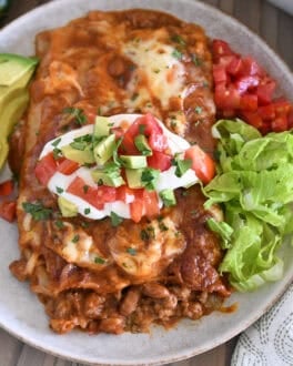 Top down view of two enchiladas on gray plate topped with red enchilada sauce, cheese, cilantro, sour cream, tomatoes, and avocados.