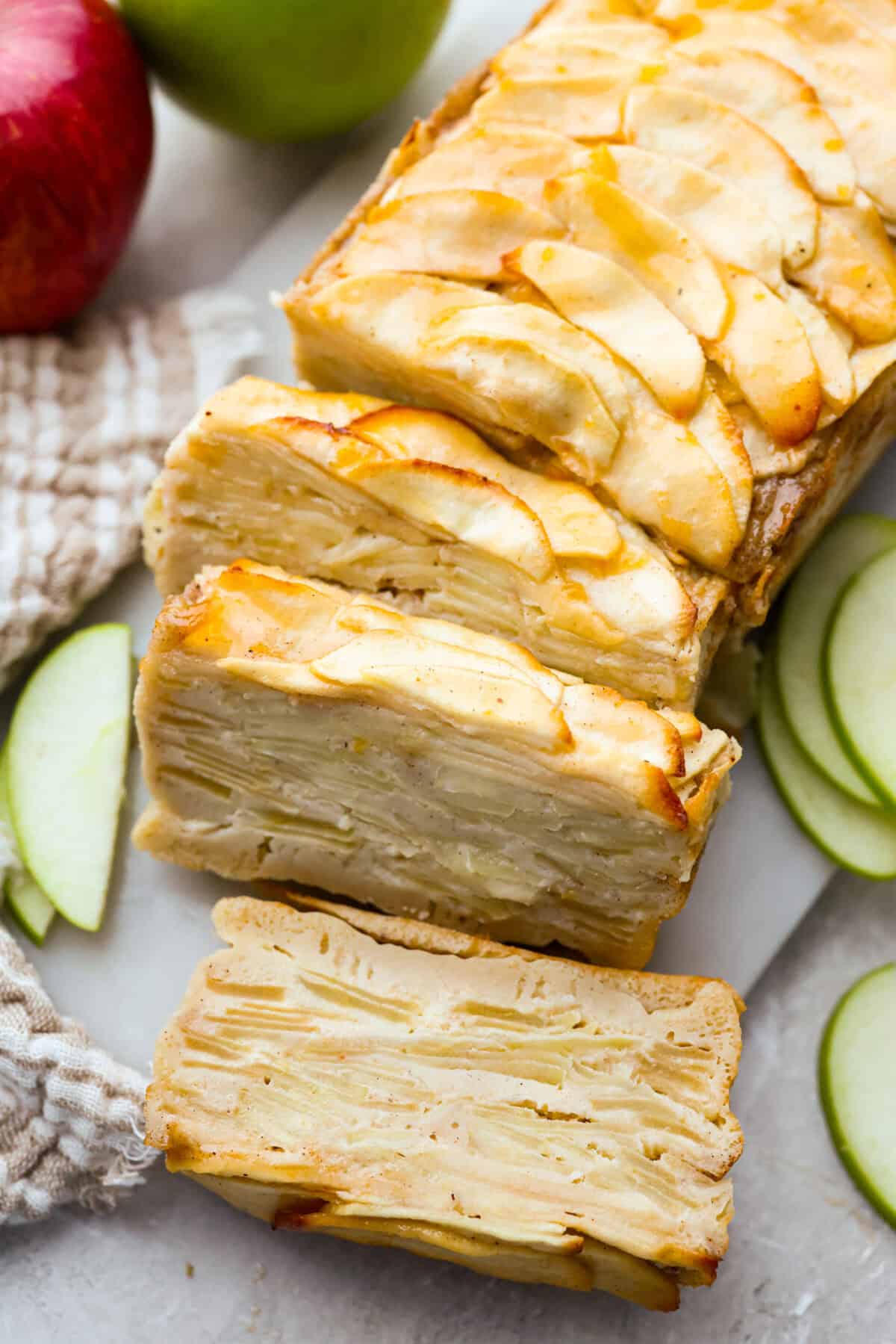Invisible Apple Cake with a few slices cut out of it on a cutting board surrounded by apples. 