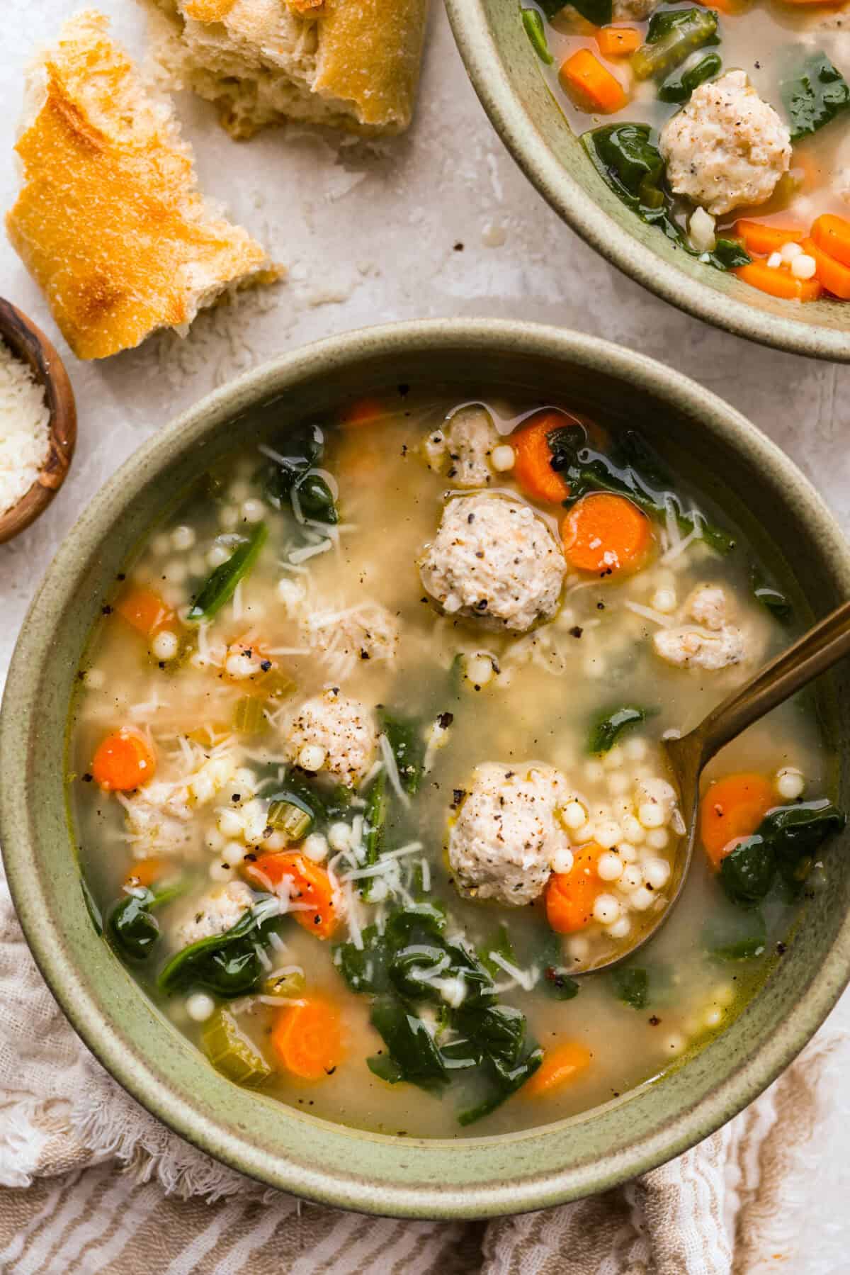 Serving bowl of Italian wedding soup surrounded by crusty bread and parmesan cheese. 