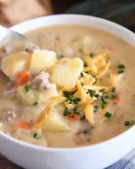 White bowl with spoon scooping ingredients in creamy cheesy soup with cooked potatoes, ground beef, and carrots and topped with shredded cheddar cheese and chopped chives.