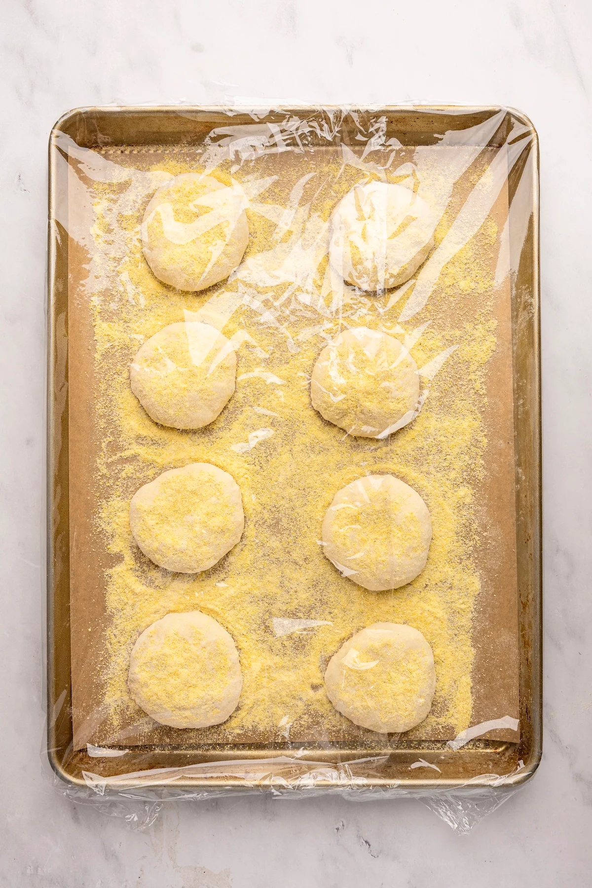 Parchment-lined baking sheet with plastic wrap over English muffin rounds 
